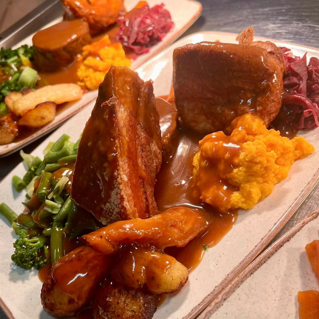 A plate of roast pork with crackling, roast potatoes, greens, carrot and swede mash, red cabbage and gravy