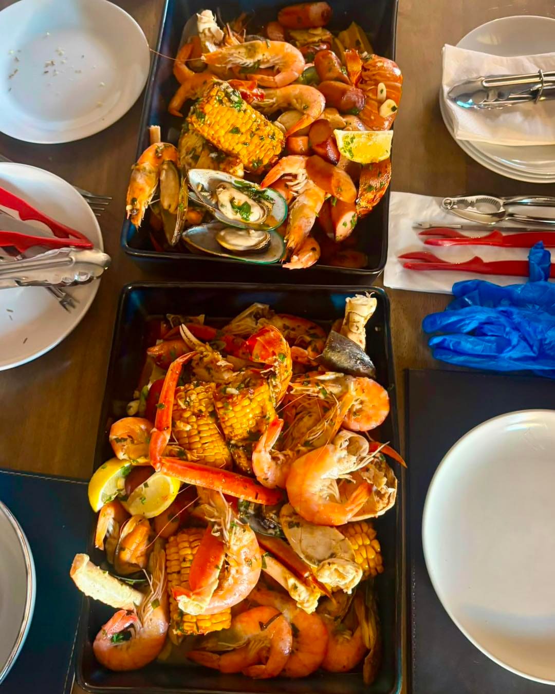 Two black dishes of seafood boil with prawns, mussels, crab legs and corn on a table