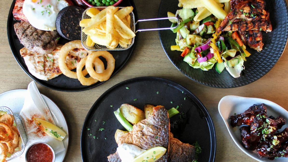 Dishes seen from above on a wooden table: a mixed grill, a basket of chips, onion rings, pan-fried fish and glazed ribs