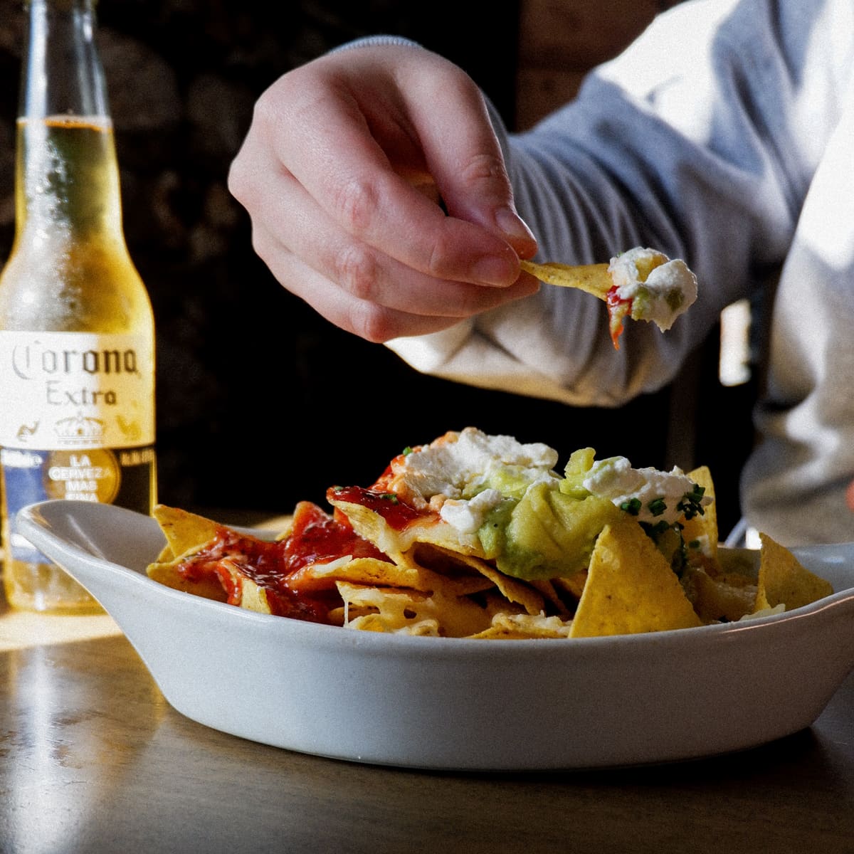Nachos with salsa, guacamole and sour cream next to a bottle of Corona, with a hand holding a loaded chip