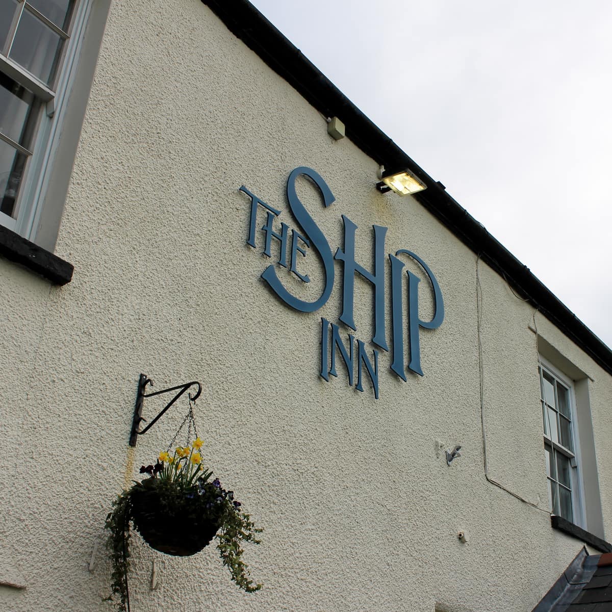 Blue “THE SHIP INN” lettering on the pub wall above a hanging flower basket